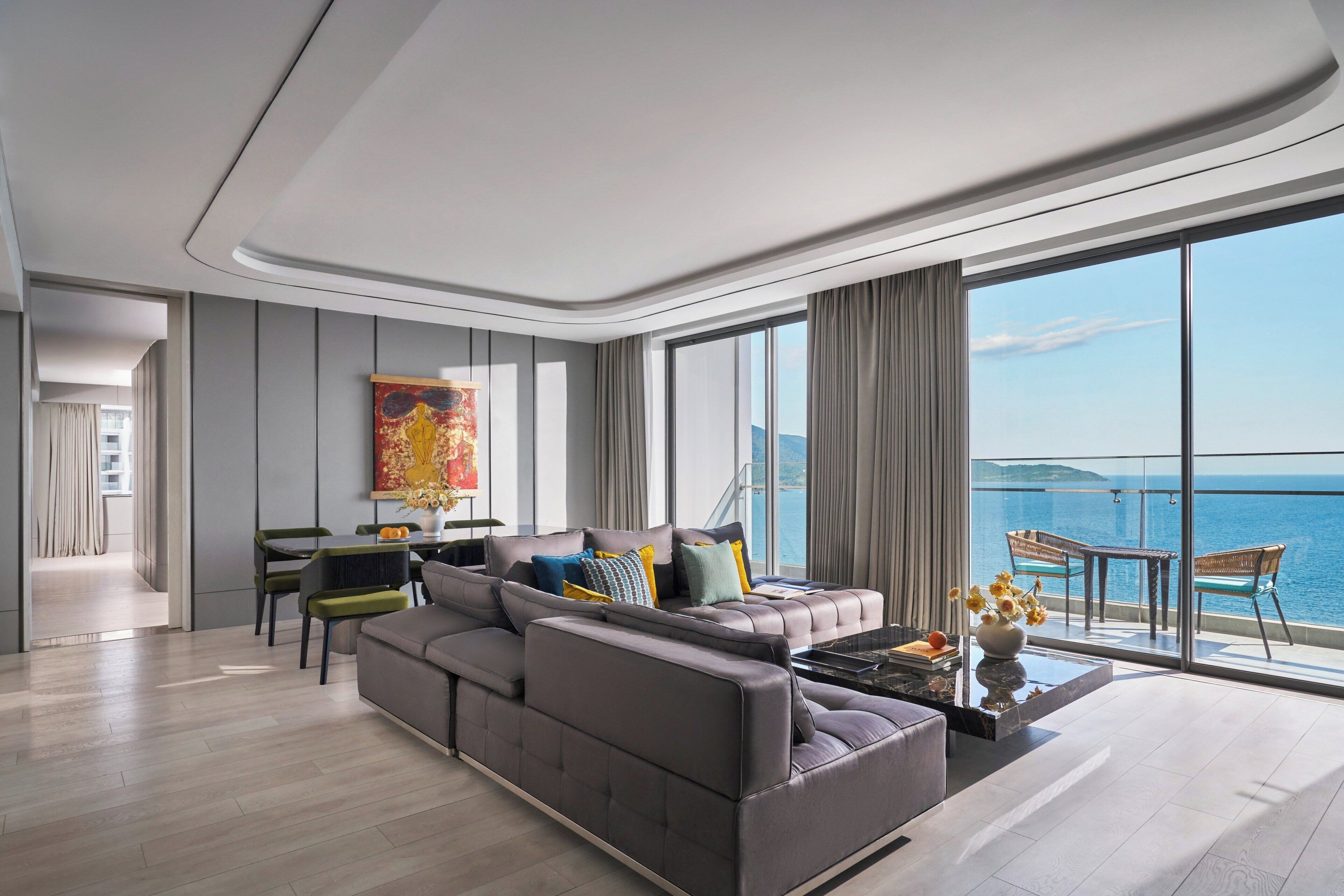 Una sala de estar moderna con un amplio sofá seccional gris, una zona de comedor con mesa y sillas, y un gran ventanal con vistas al mar.