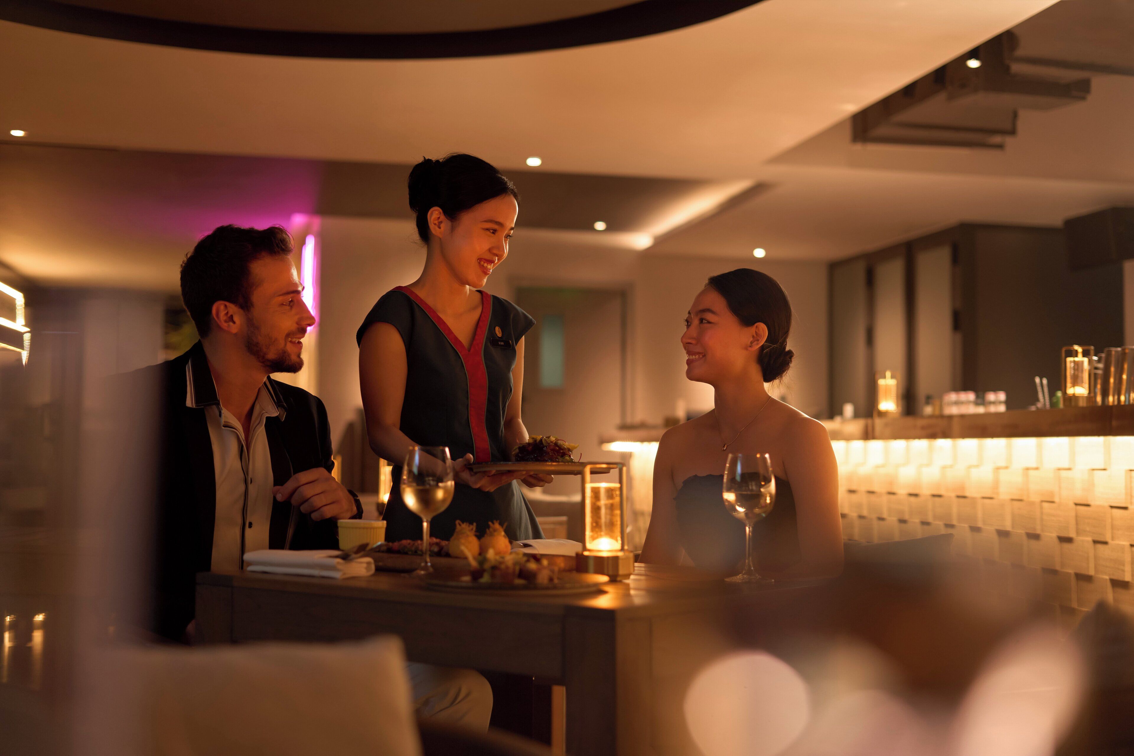 A modern dining area with a bar, a waiter serving food, and guests seated at a table with wine glasses.