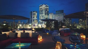 A rooftop lounge with outdoor seating, city skyline views, and illuminated tables.