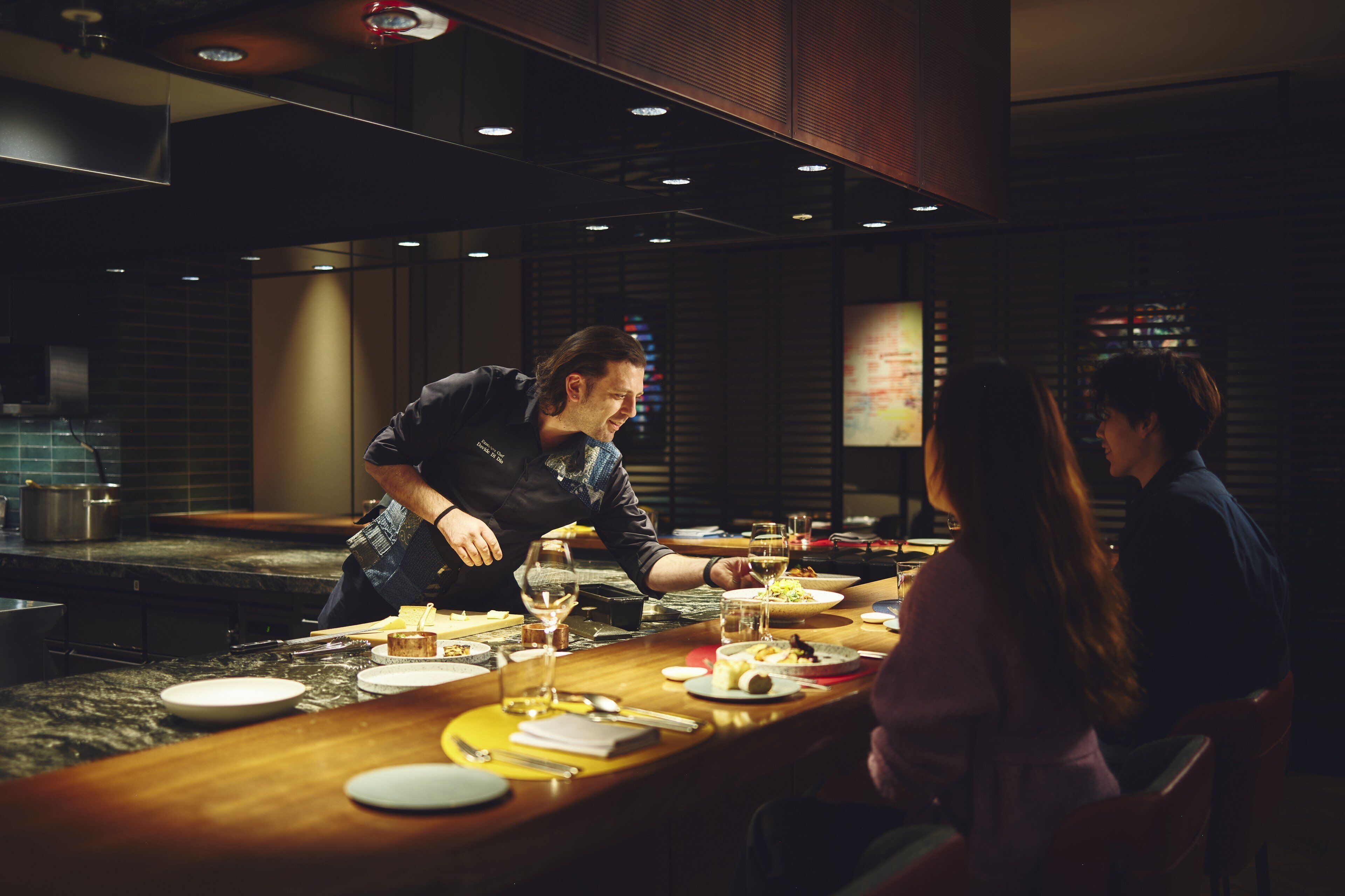 Un restaurant à la lumière tamisée, avec un bar, un chef qui prépare les plats et des clients assis à des tables.