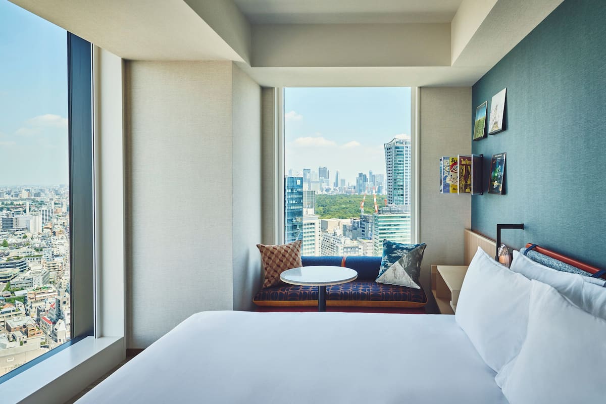 A modern hotel room with a large bed, a small round table, and a view of the cityscape through a large window.