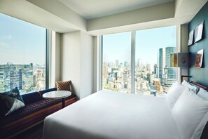Habitación de hotel con una cama grande, una mesita redonda y un sofá que ofrece vistas a un paisaje urbano a través de amplias ventanas.
