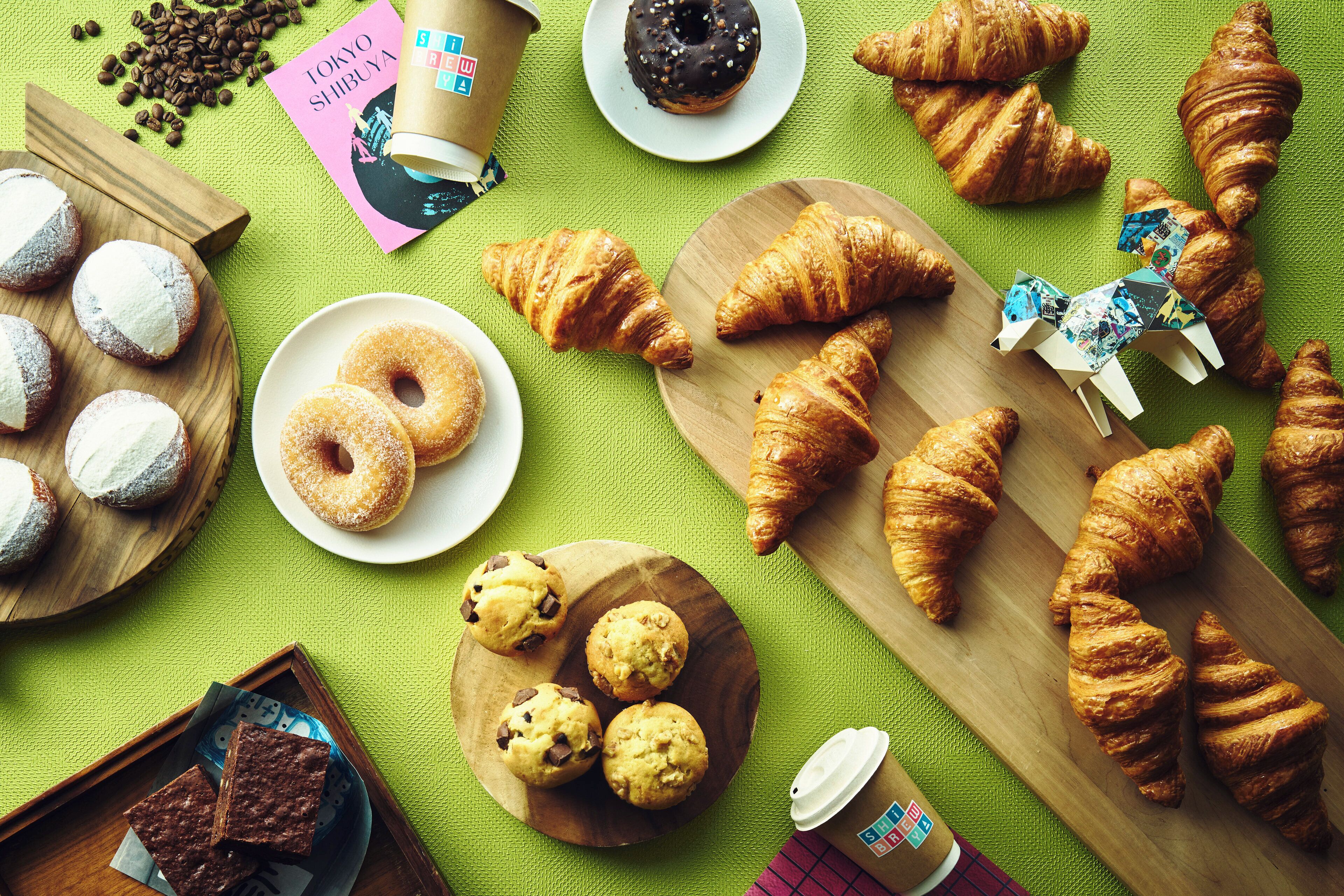 A variety of pastries and baked goods on wooden boards and plates, with a coffee cup and a travel magazine on a green textured surface.