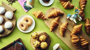 A variety of pastries and baked goods on wooden boards and plates, with a coffee cup and a travel magazine on a green textured surface.