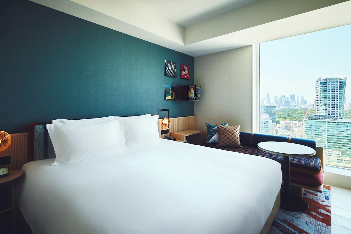 A hotel room with a large bed, a sofa, a small table, and a view of the cityscape.
