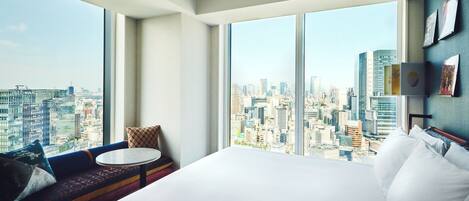 A hotel room with a large bed, a small round table, and a sofa with a view of the cityscape.