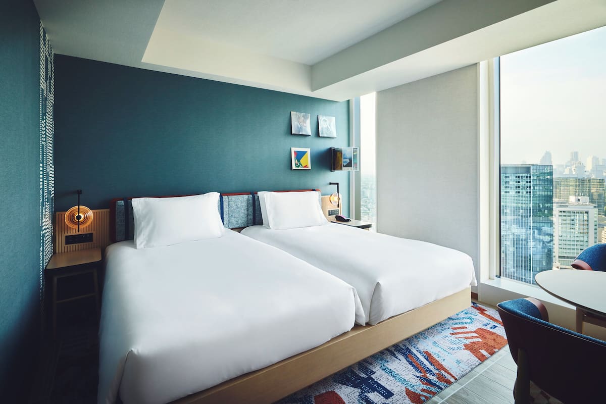 A modern hotel room with two beds, a city view, and a small dining area.