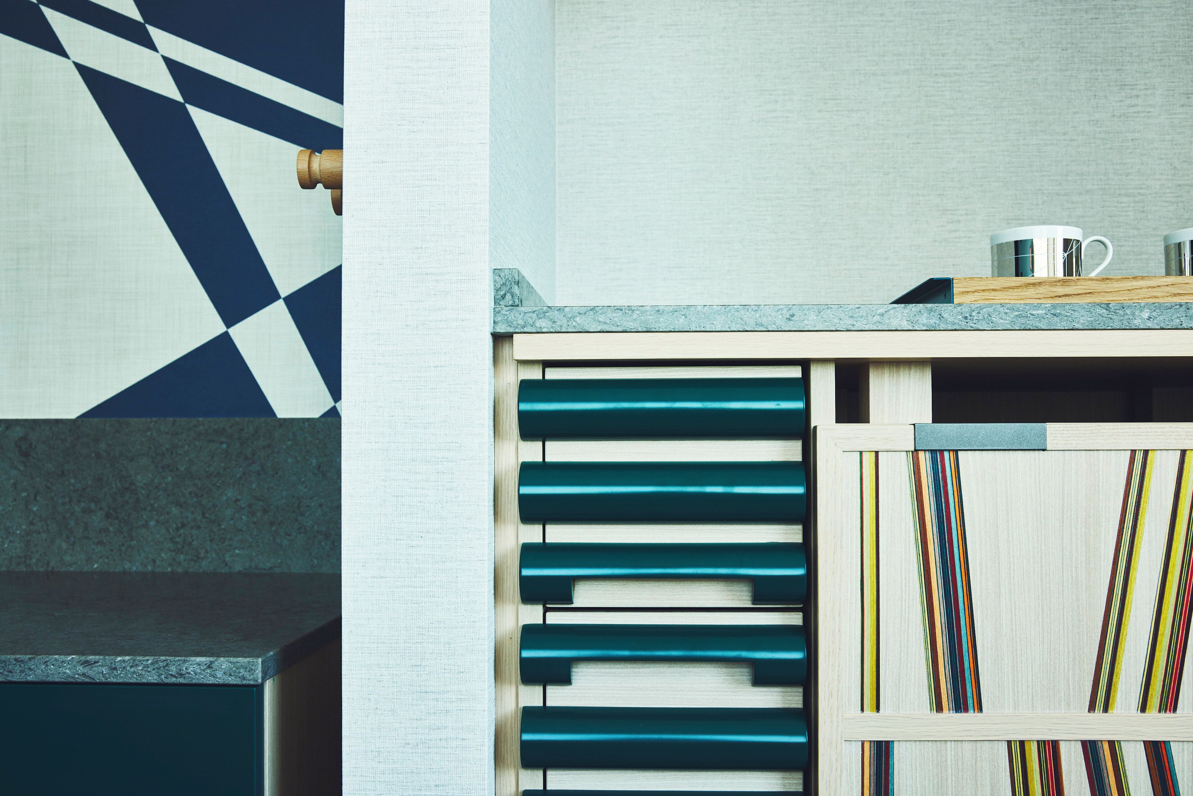 A kitchen with a geometric patterned wall, a wooden cabinet with drawers, and a countertop with a kettle and a mug.