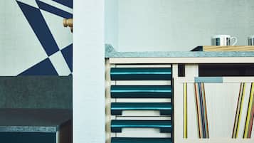 A kitchen with a geometric patterned wall, a wooden cabinet with drawers, and a countertop with a kettle and a mug.