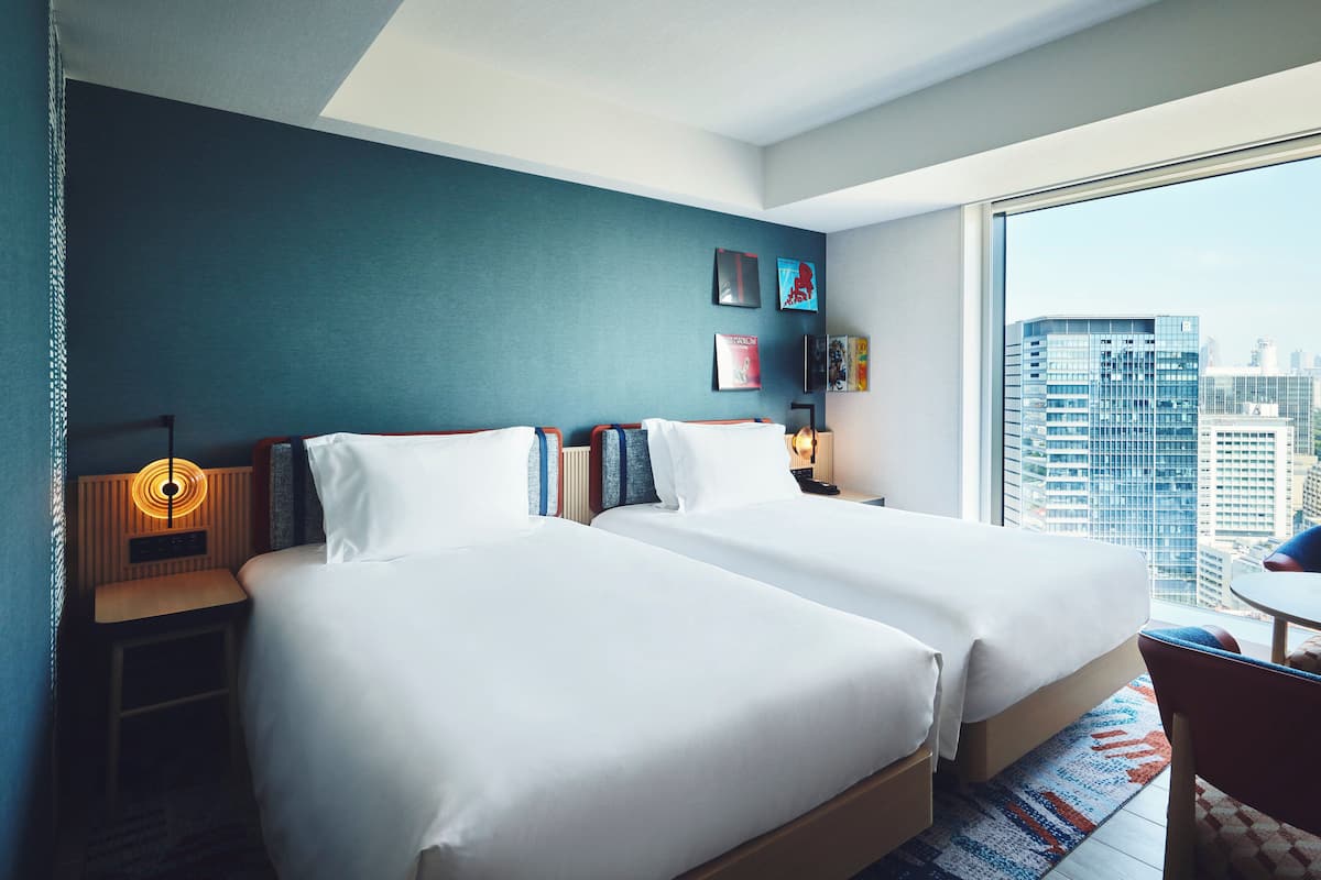 Habitación de hotel con dos camas, una mesita de noche, una lámpara y vista a la ciudad.
