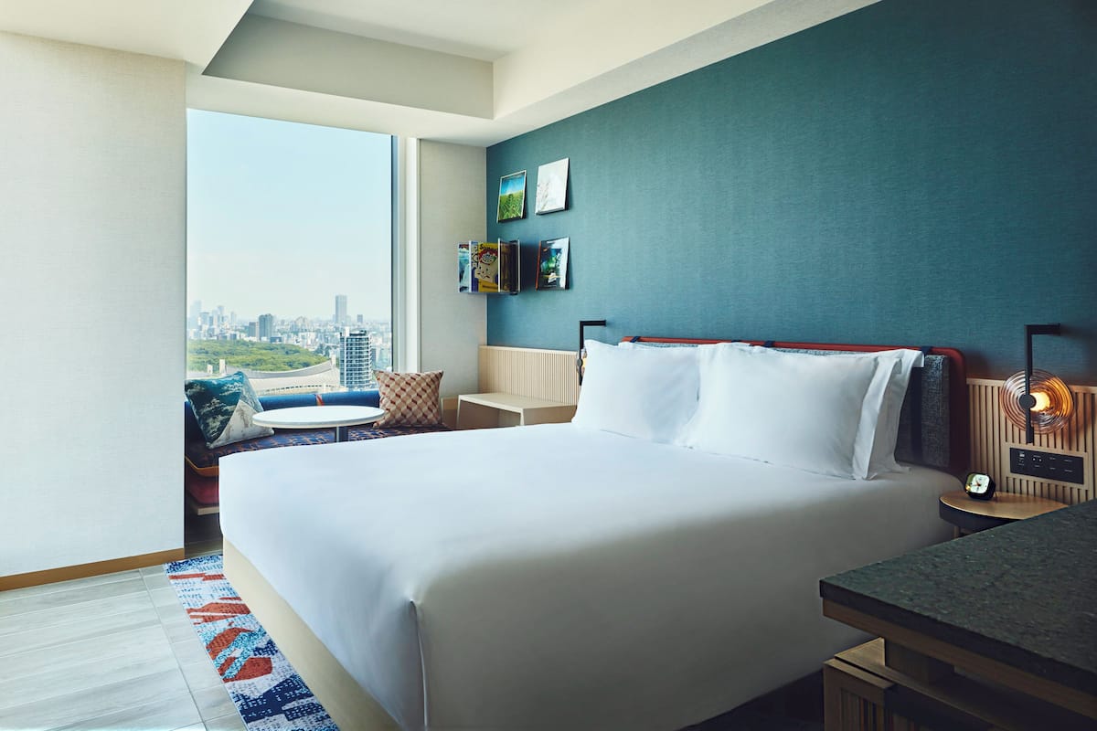 A modern hotel room with a large bed, a small table, a chair, and a view of the city.
