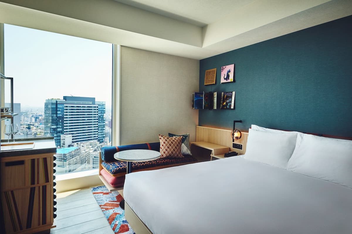 A hotel room with a large bed, a seating area with a view of the city, a small table, and a bookshelf.