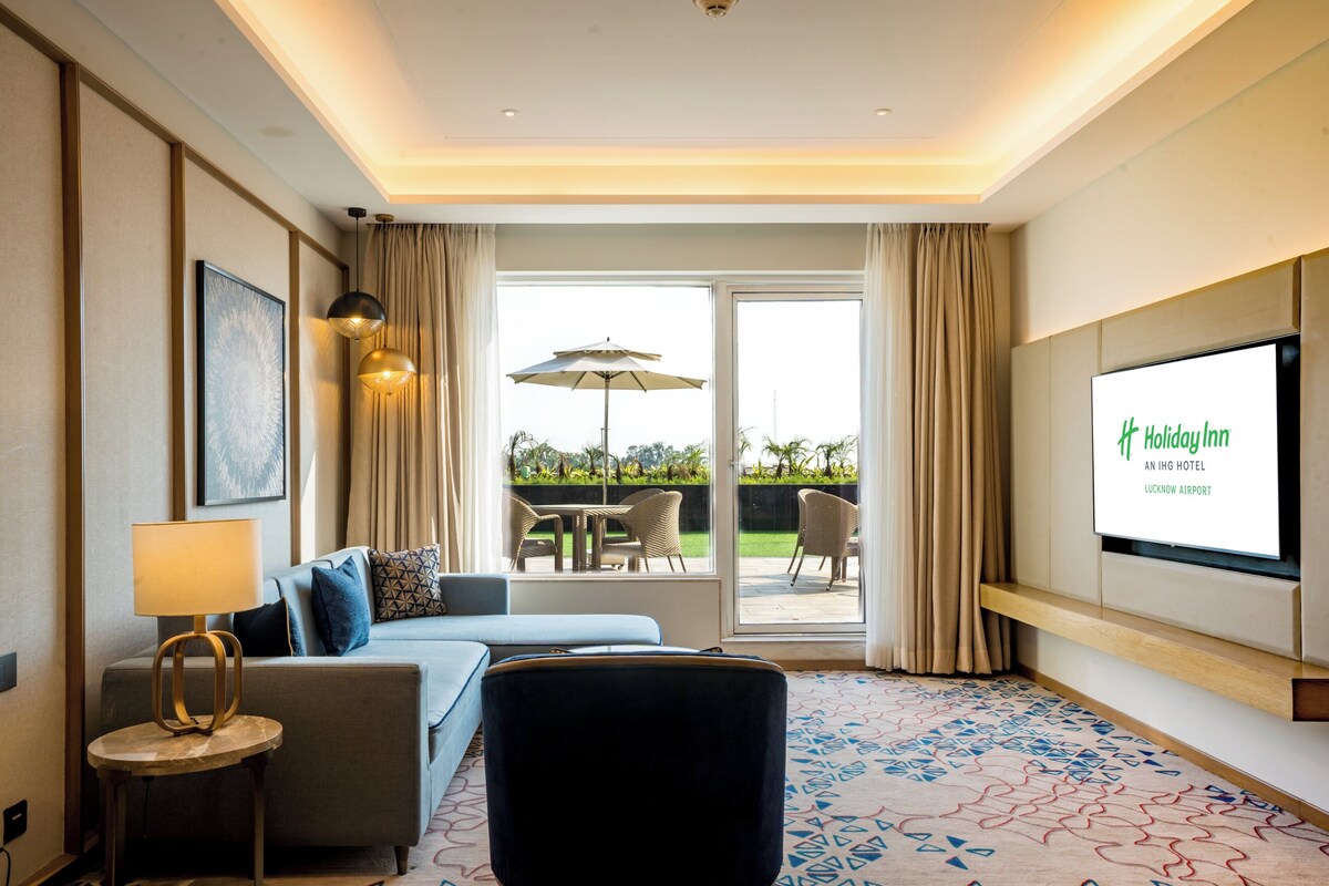 Een hotelkamer met een bank, een klein tafeltje met een lamp, een groot televisiescherm waarop de naam van het hotel staat, en een uitzicht op het buitengebied door een glazen deur.