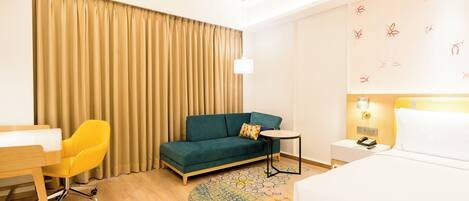 A hotel room with a green sofa, a yellow chair, a small round table, a bed, a desk, and a patterned rug.