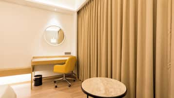 Habitación de hotel con un escritorio de madera, una silla amarilla, una mesa redonda, una alfombra pequeña y cortinas color beige.
