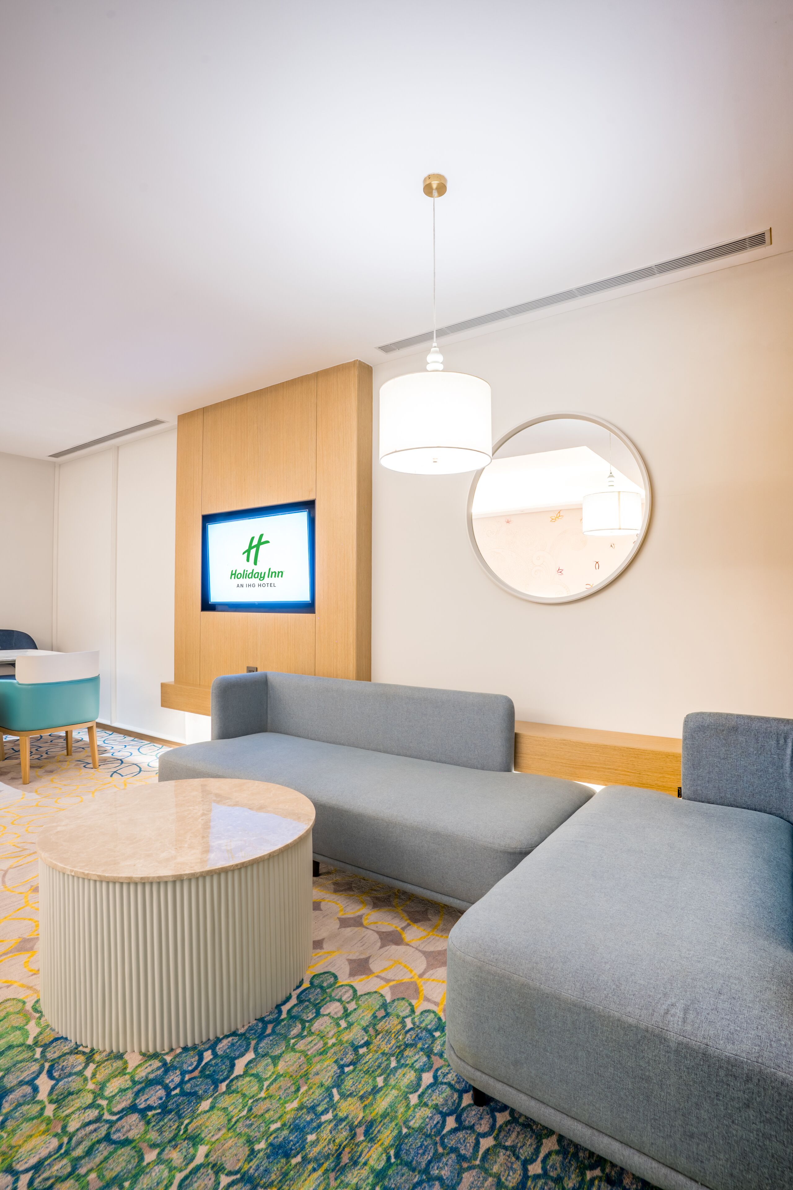 A modern hotel lobby with a seating area, a television displaying the hotel name, and a round mirror on the wall.