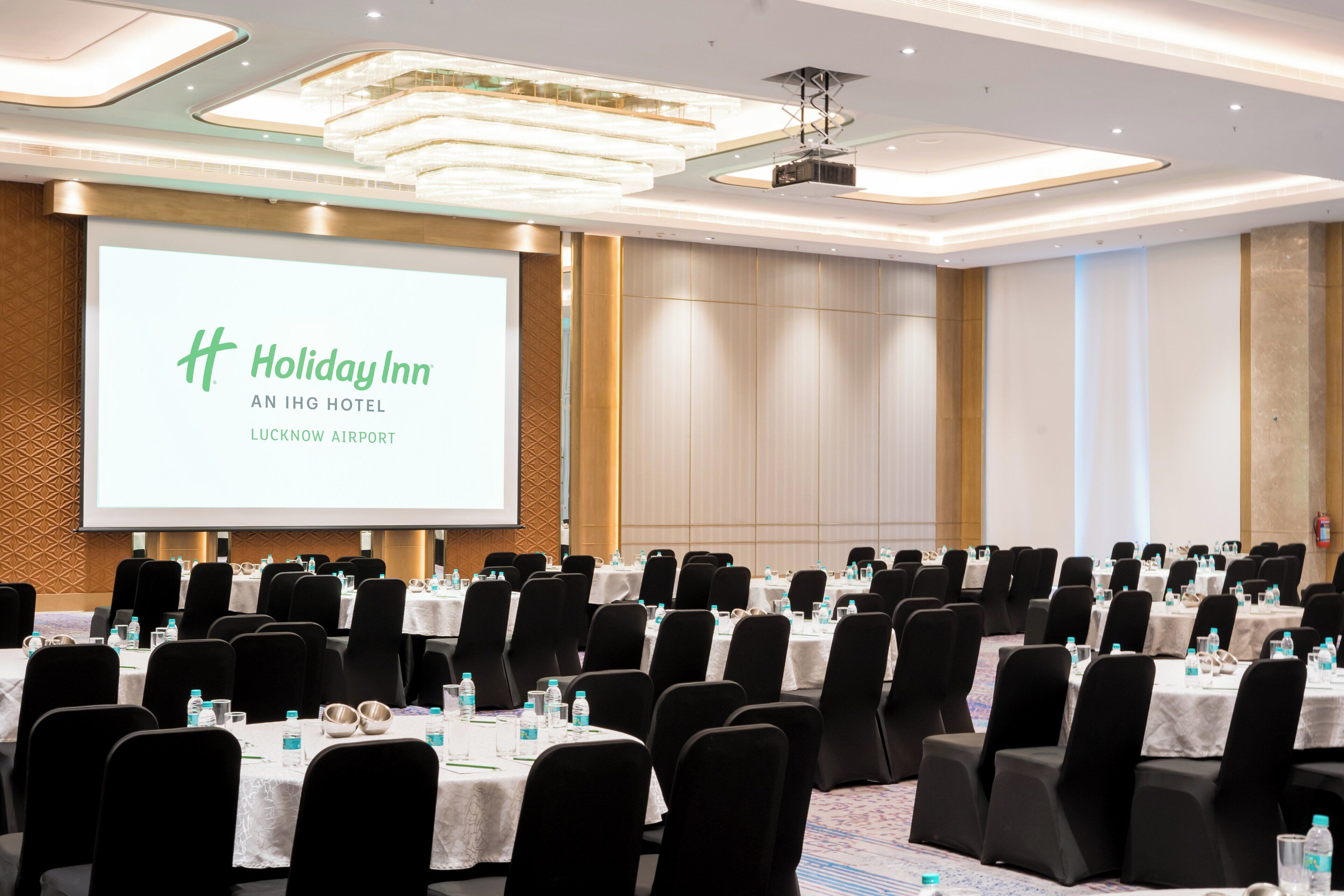 A conference room at Holiday Inn with a large screen displaying the hotel name, arranged tables with chairs, and a carpeted floor.
