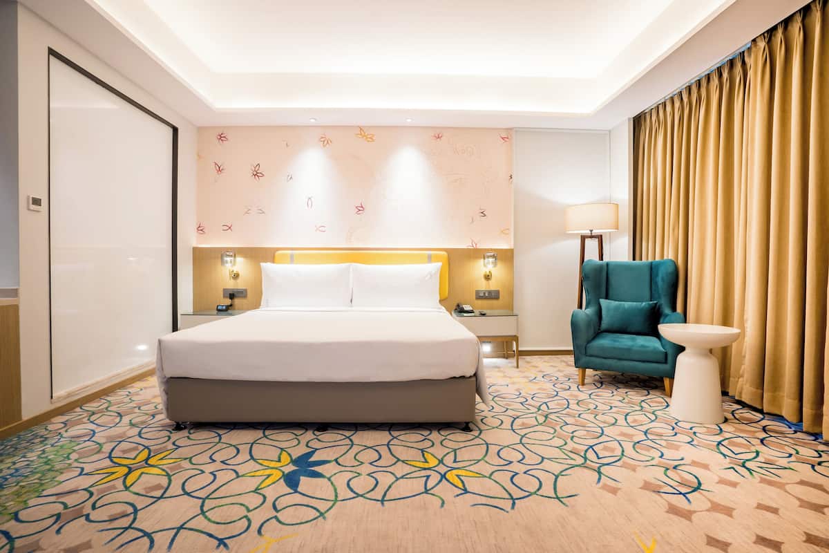 Ein Hotelzimmer mit einem großen Bett, einem türkisfarbenen Sessel, einem kleinen weißen Tisch und einem gemusterten Teppich.