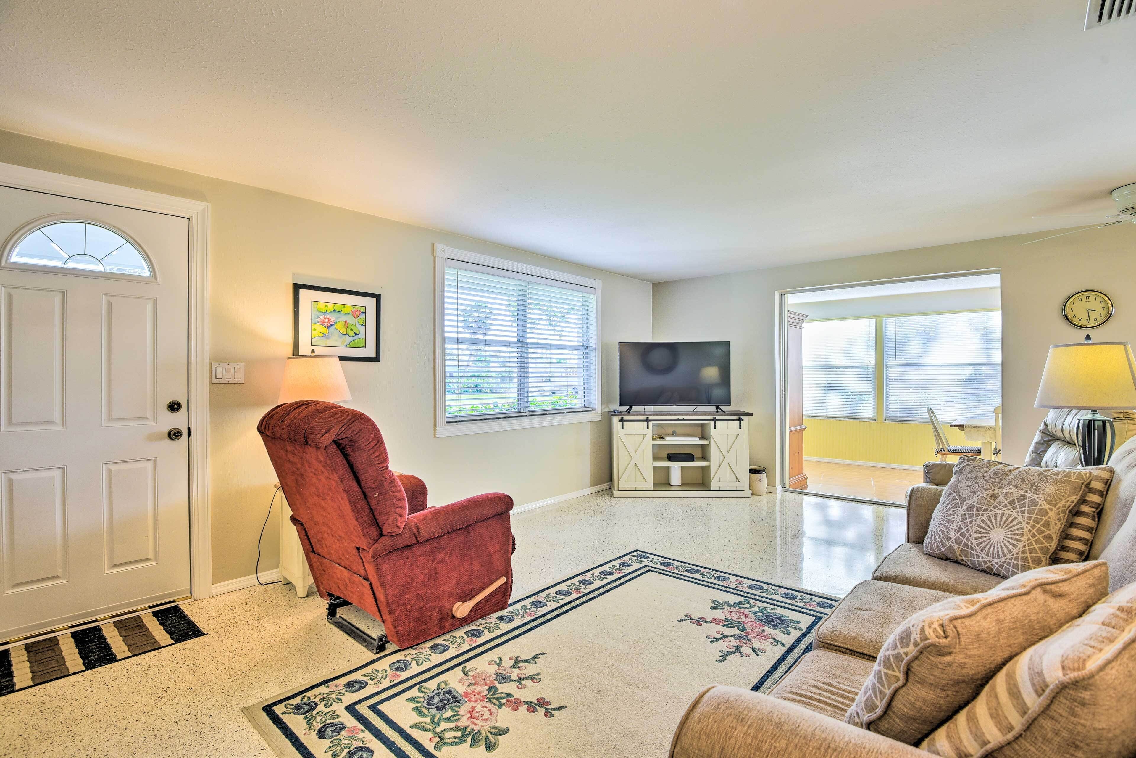 A living room with a red armchair, a beige sofa, a TV on a white stand, and a large window with blinds.