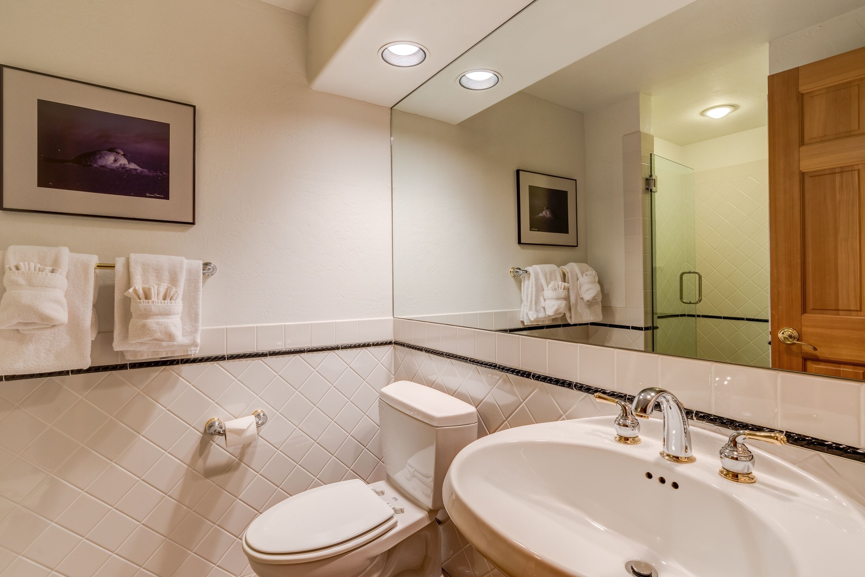 A bathroom with a toilet, sink, and mirror. There are towels hanging on the wall and a framed picture above the toilet.