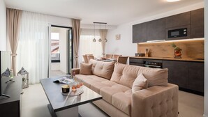 A modern living room with a beige sofa, a dining table, and a kitchen area with dark cabinets and a microwave. - Beach Villa Valentina Apartment IV (Okrug Gornji)