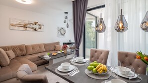 Un salon moderne avec un canapé, une table à manger et des chaises. Les murs sont ornés de tableaux abstraits, il y a une fenêtre avec des rideaux et des lampes suspendues au-dessus de la table.