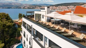 A modern building with a rooftop pool area, including lounge chairs and umbrellas, overlooking a coastal town and a body of water. - Beach Villa Valentina Apartment IX (Okrug Gornji)