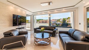 A modern living room with a flat-screen TV mounted on a textured wall, leather sofas, a glass coffee table, and a large window offering a city view.