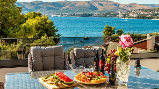 A table with a fruit platter, wine bottles, and a vase with flowers, overlooking a coastal view with mountains and boats.