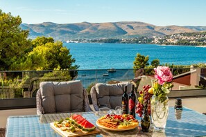 Een tafel met een fruitplateau, wijnflessen en een vaas met bloemen, met uitzicht op de kust, bergen en boten.