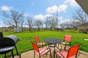 Un patio exterior con una mesa redonda y sillas, rodeado por un área de césped con árboles y cielo despejado.