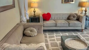 A living room with a beige sofa, a smaller sofa, and a patterned rug. There are two lamps on side tables, a red heart-shaped pillow, and a decorative tray on the floor. - LemonTree Cottage firepit and hottub, 8 minutes from the beach (New Smyrna Beach)