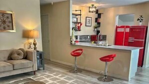 A modern living room with a sofa, a coffee table, and two red bar stools. A kitchen area with a red refrigerator and a bar counter is visible in the background. - LemonTree Cottage firepit and hottub, 8 minutes from the beach (New Smyrna Beach)