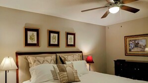 Ein Schlafzimmer mit einem Bett, einem Deckenventilator, gerahmten Bildern an der Wand und einem Nachttisch mit einer Lampe.