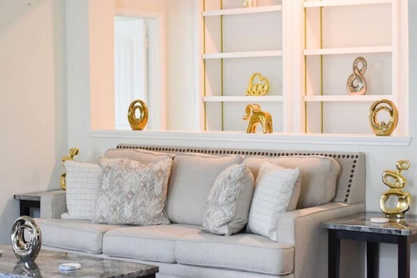 Un salon avec un canapé beige, une table basse en marbre et un mur blanc orné d’étagères où sont disposés des objets décoratifs.