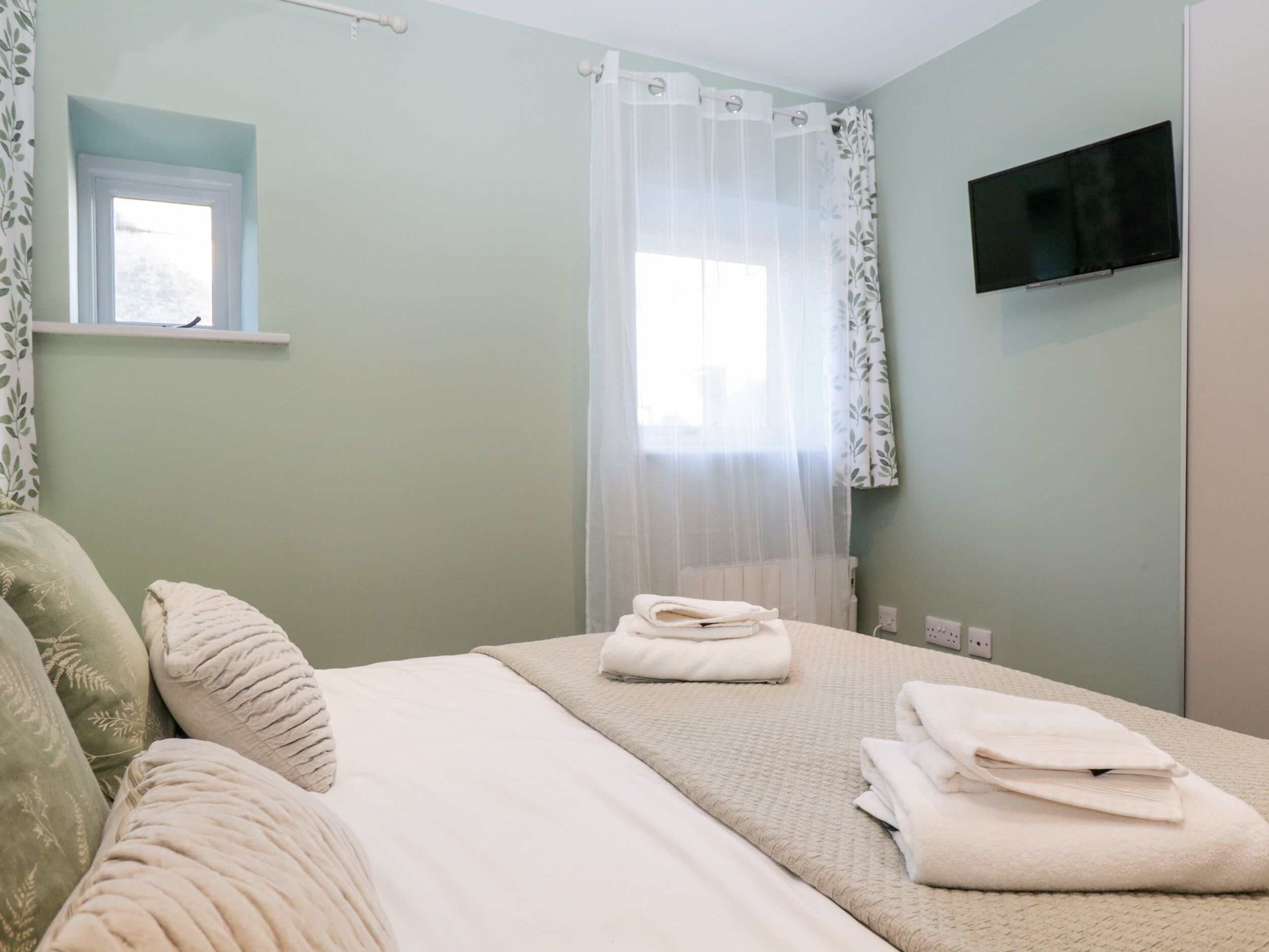 A neatly arranged bedroom with a bed, pillows, a TV mounted on the wall, and a window with sheer curtains.