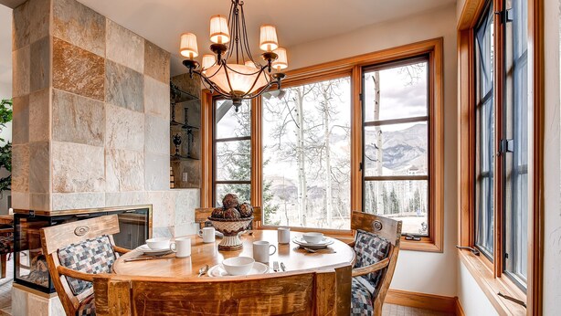 Un coin repas avec une table en bois, des chaises et un lustre. De grandes fenêtres offrent une vue sur les arbres et les montagnes.