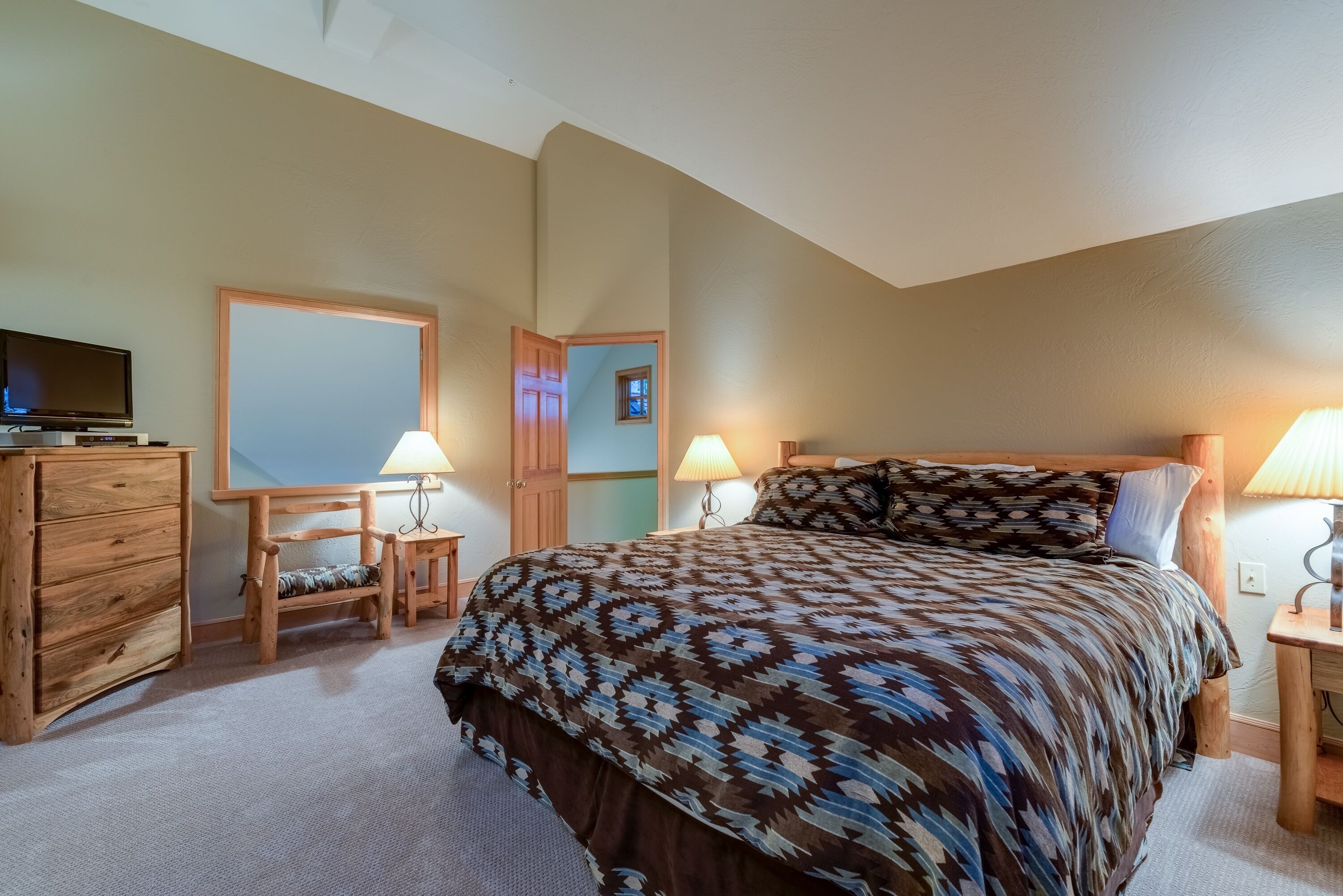 A bedroom with a bed, wooden dresser, two nightstands with lamps, a television, and a mirror.