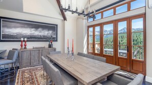 A spacious dining area with a large wooden table, grey upholstered chairs, and a view of mountains through large windows. - Slopeside Sanctuary Penthouse 3 Bedroom Condo by Alpine Lodging Telluride (Telluride)