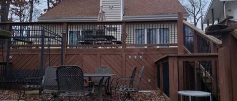 Una terraza de madera con sillas y una mesa de metal, un patio trasero cercado y una casa con chimenea.