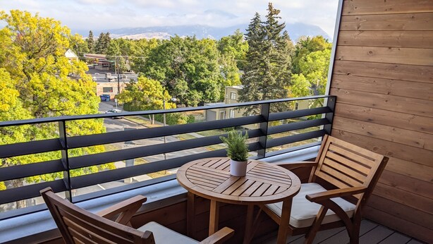 A wooden deck with a round table and two chairs, a potted plant, and a view of trees and buildings. - Bozeman Summer Getaway: Climate-Controlled Garage + Mountain Views! (Bozeman)