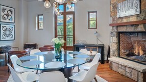 A dining area with a glass table surrounded by white chairs, a chandelier, and a stone fireplace. - Mt Wilson Basecamp 4 Bedroom Condo by Alpine Lodging Telluride (Telluride)