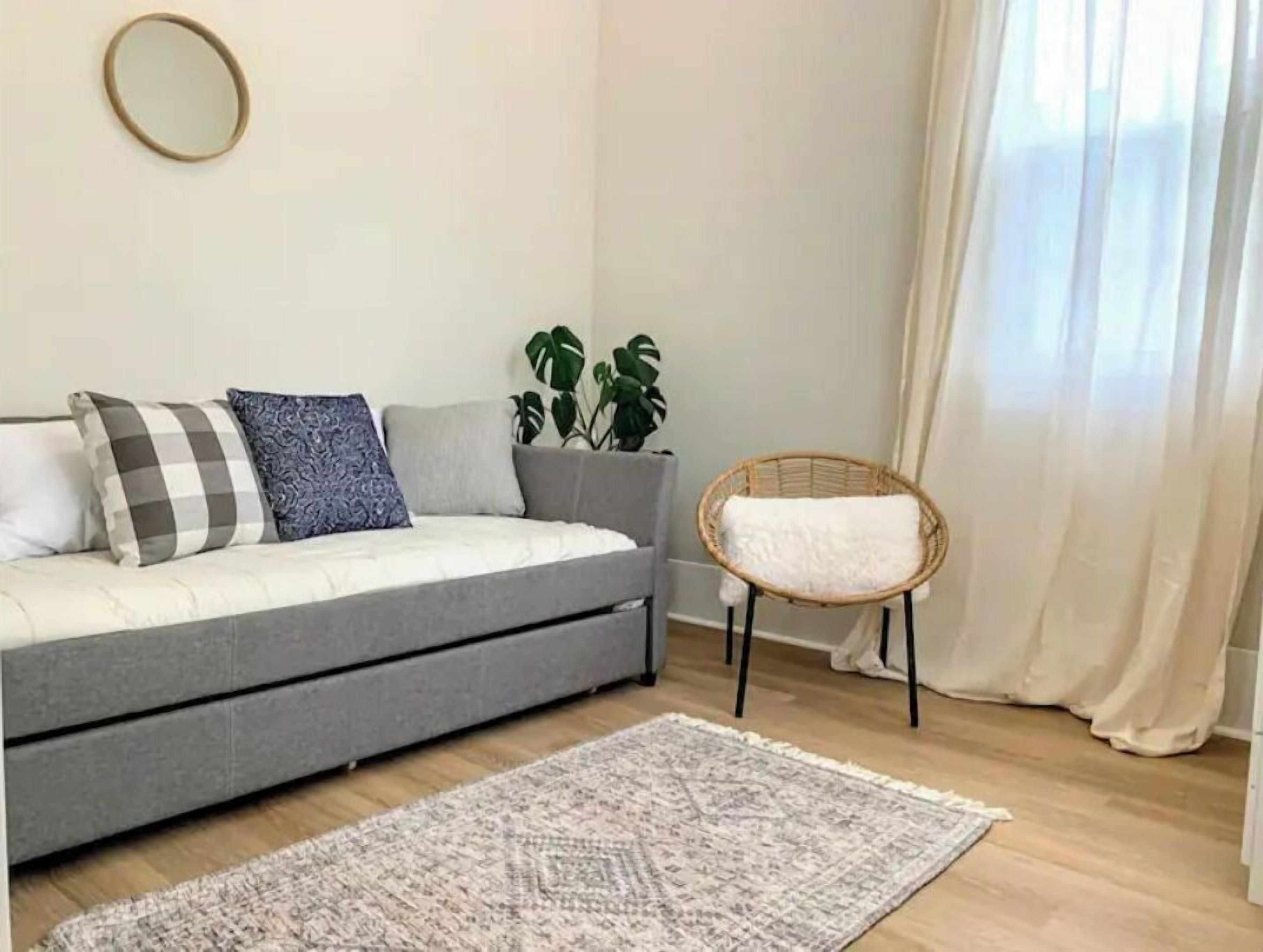 A modern living room with a grey sofa, patterned cushions, a wicker chair, a round mirror, and a potted plant.