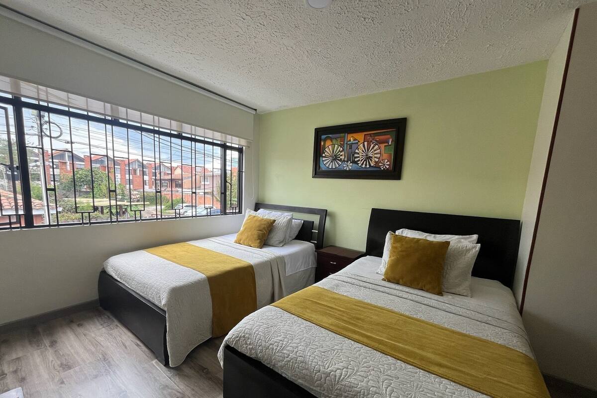Een hotelkamer met twee bedden, een raam met tralies, een schilderij aan de muur en uitzicht op gebouwen.