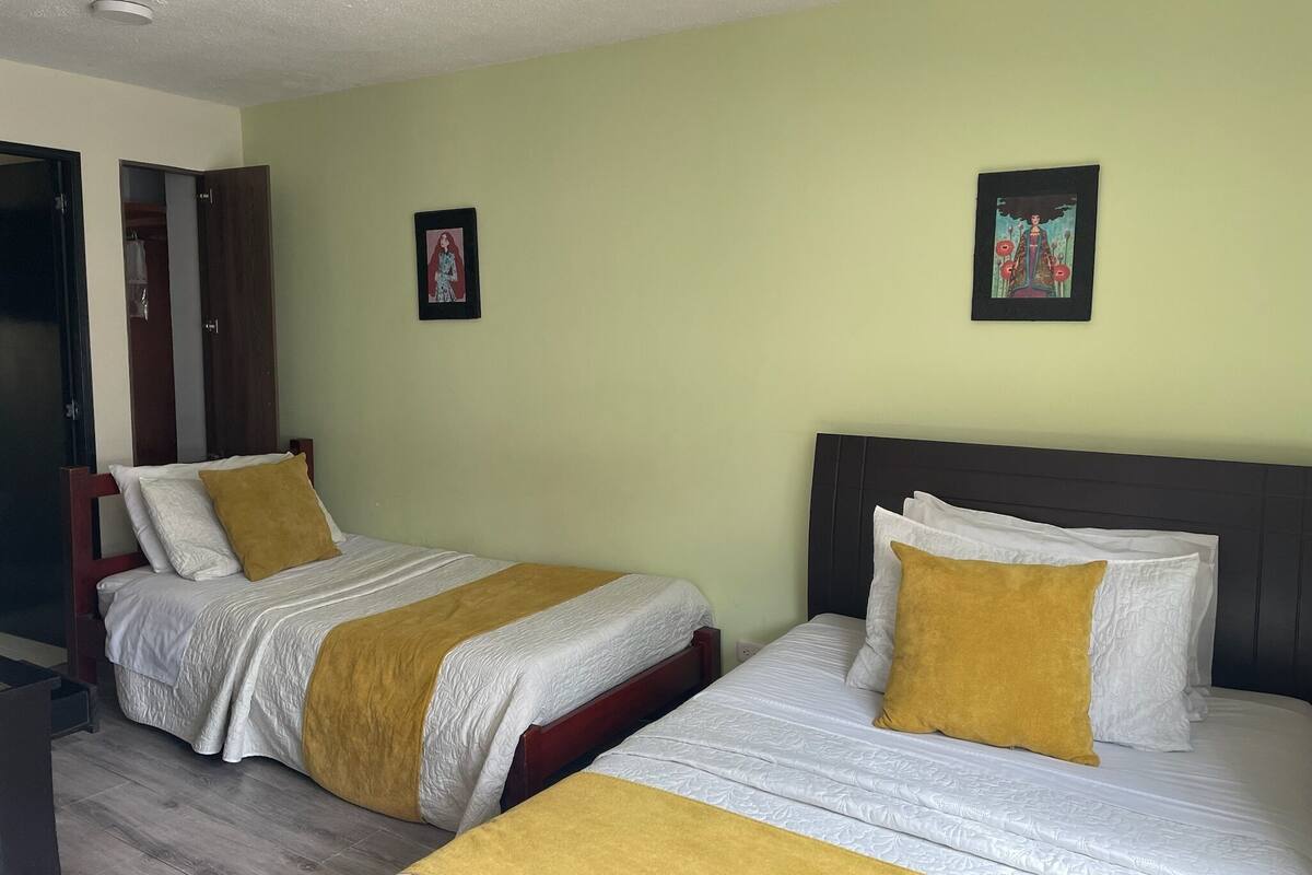 Een hotelkamer met twee bedden, elk voorzien van witte beddengoed en een mosterdgele plaid. De kamer heeft lichtgroene muren, houten hoofdborden en twee ingelijste foto’s aan de wand.