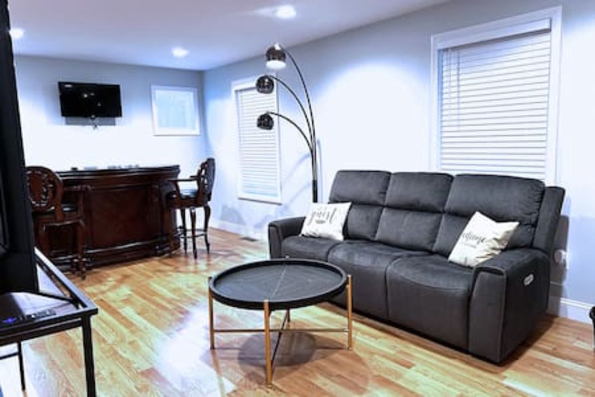 A living room with a sofa, a round coffee table, a desk with a chair, a television mounted on the wall, and a window with blinds.