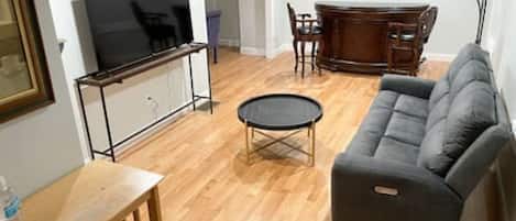 A living room with a flat-screen TV, a gray sofa, a wooden coffee table, and a dining area with a wooden table and chairs.