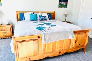 Una cama de madera con colcha blanca y azul estampada, una mesita de noche de madera con una lámpara, y una puerta blanca en una habitación con piso alfombrado.