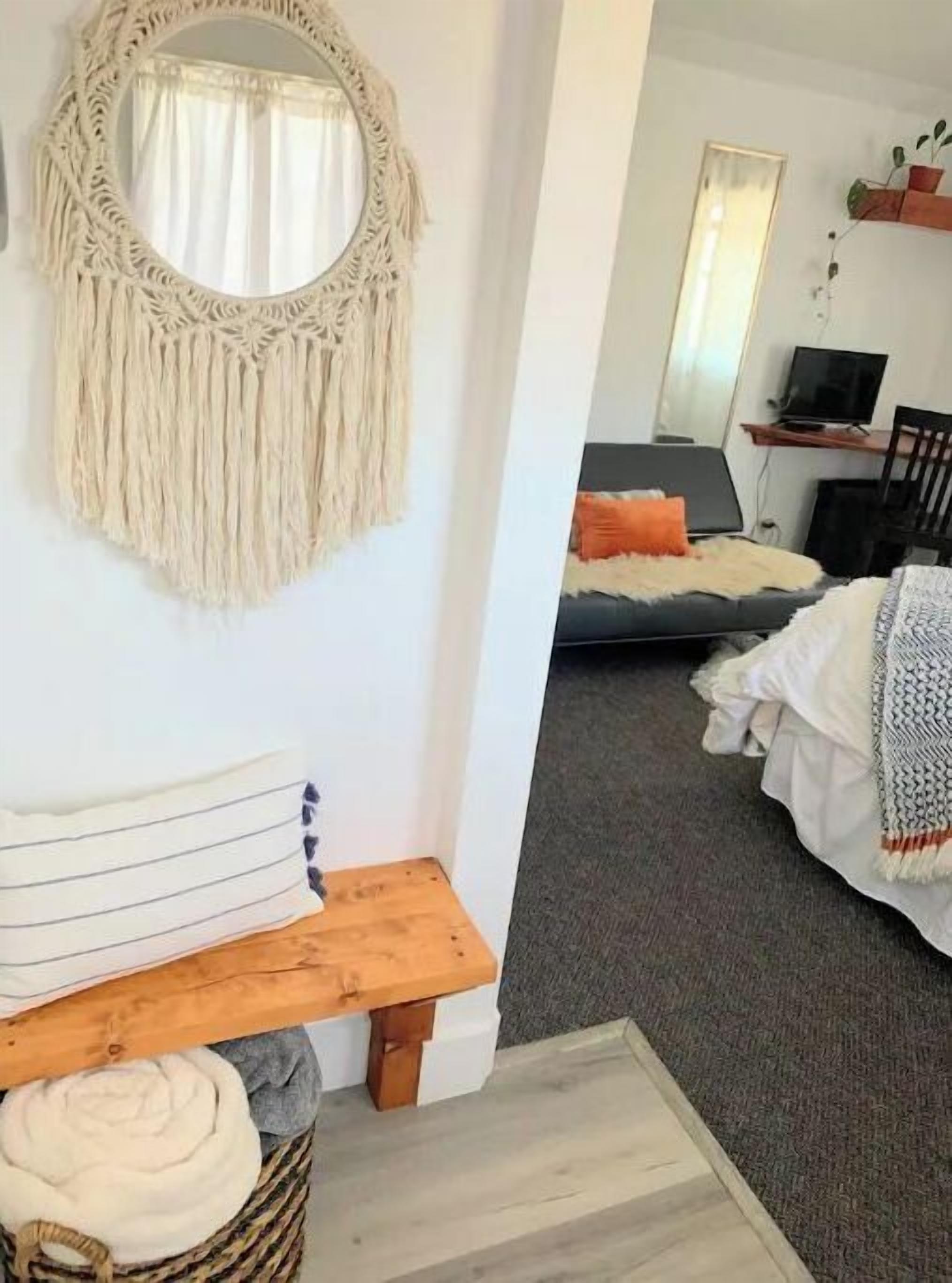 Una habitación acogedora con un tapiz de macramé en la pared, un banco de madera y una cama con una manta gris y un cojín naranja.