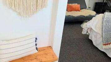 Una habitación acogedora con un tapiz de macramé en la pared, un banco de madera y una cama con una manta gris y un cojín naranja.